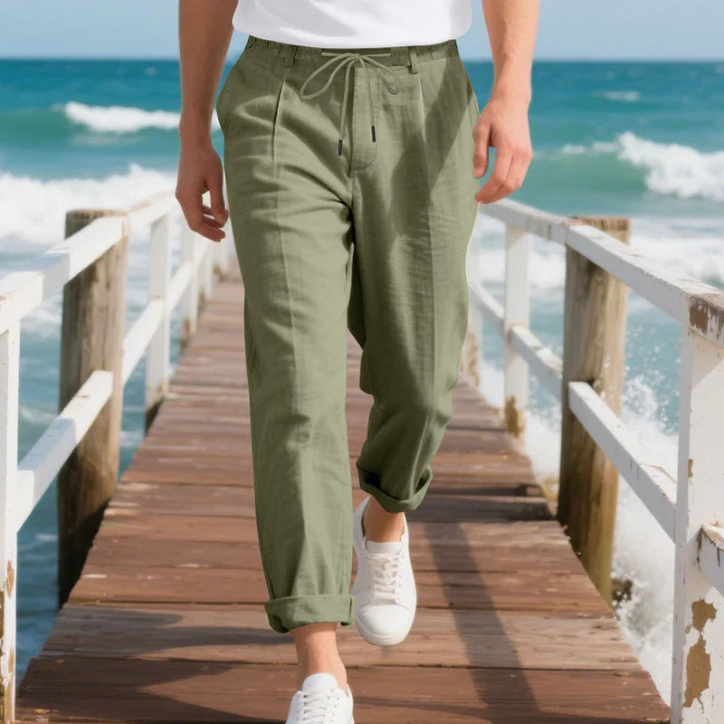 Man wearing olive green drawstring pants and white sneakers walking on wooden pier by the ocean