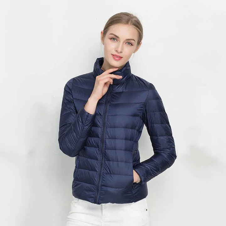 Woman wearing navy blue quilted lightweight puffer jacket and white pants against white background
