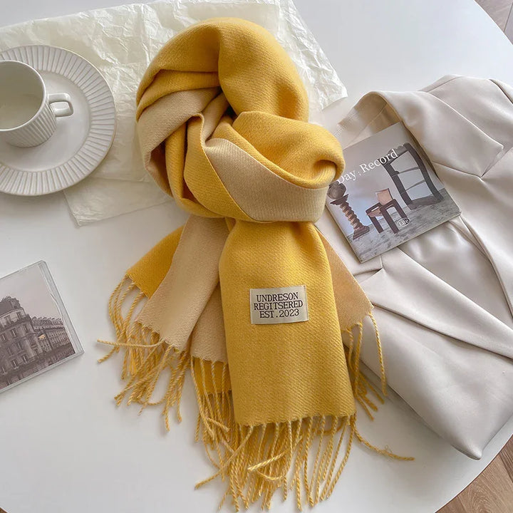 Yellow and beige fringed scarf with label on white table next to white blazer, coffee cup, and book