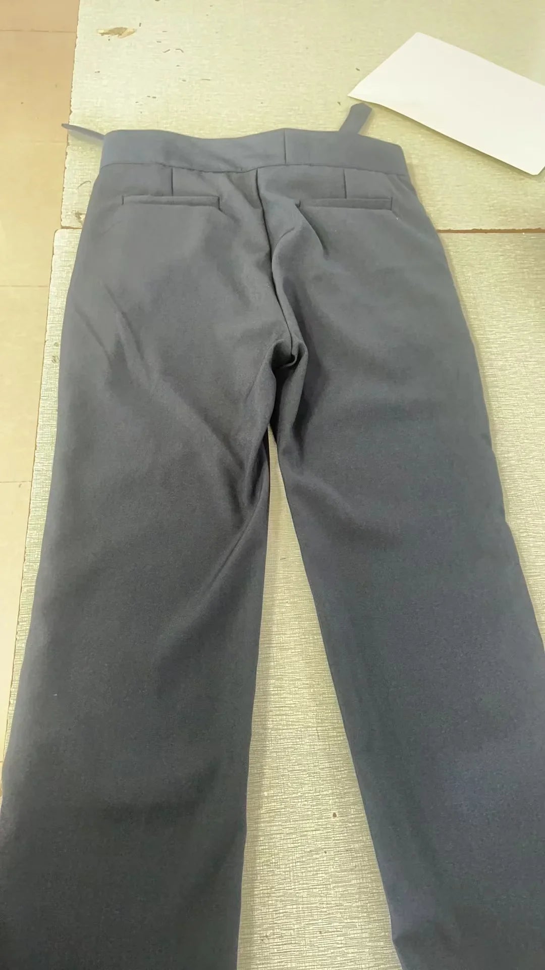 Back view of dark gray formal trousers with two back pockets on a beige textured surface