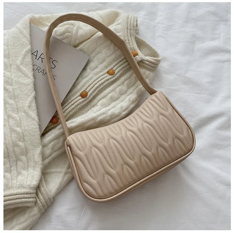 Beige quilted handbag with a long strap placed on cream cable knit sweater with orange buttons on white surface