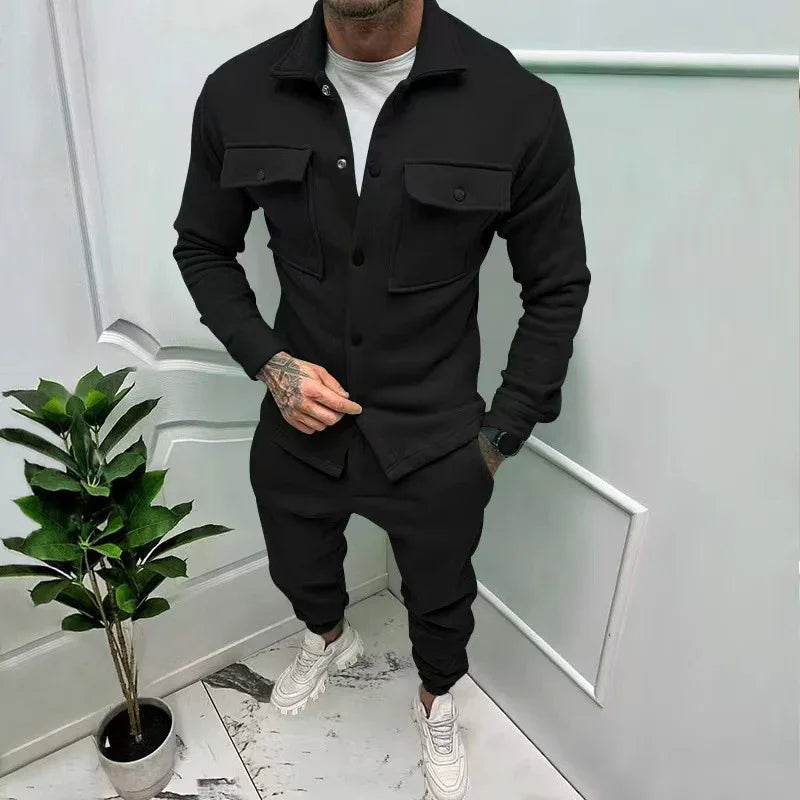 Man wearing black button-up jacket with pockets and matching joggers, standing next to a potted plant indoors