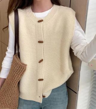 Woman wearing beige knitted button-up vest over white long sleeve shirt with blue jeans, holding brown woven tote bag