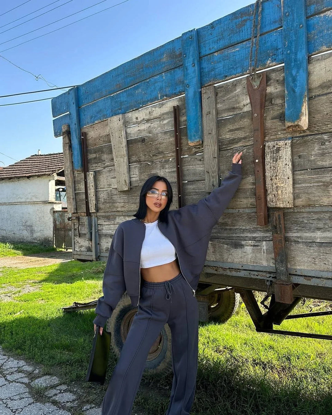 Fashionable woman in gray tracksuit and white crop top posing outdoors by rustic wooden trailer under clear blue sky