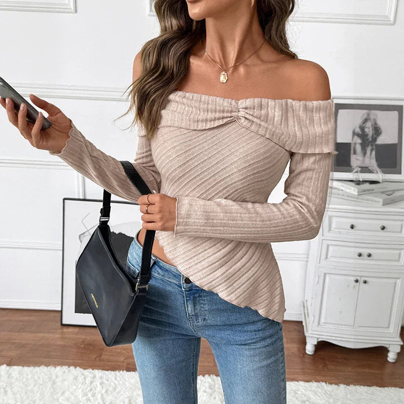 Woman wearing beige off-shoulder ribbed knit sweater with jeans, holding black shoulder bag indoors