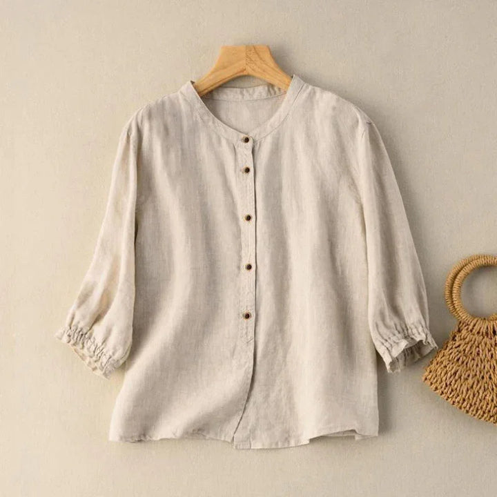 Beige long sleeve linen blouse with buttons on wooden hanger next to woven straw handbag