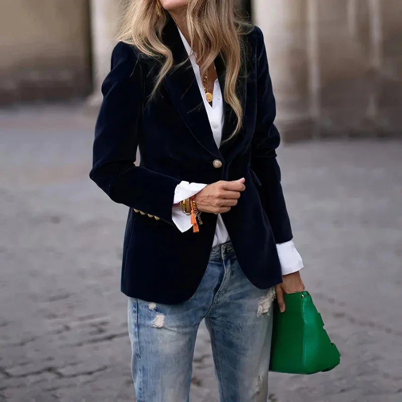 Woman wearing a navy blue velvet blazer over white shirt with ripped blue jeans, holding a green handbag