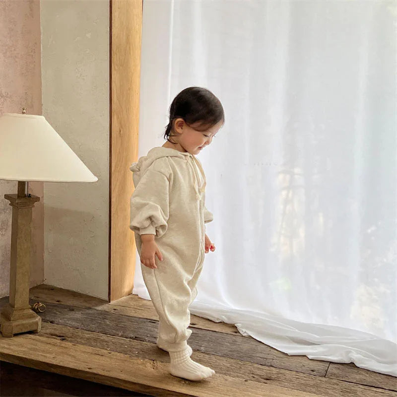 Toddler in beige hooded jumpsuit standing by sheer white curtains on wooden floor with lamp nearby