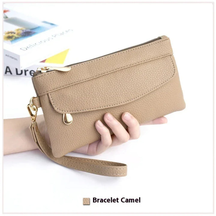Hand holding camel-colored leather wristlet clutch with gold zipper and wrist strap on white background