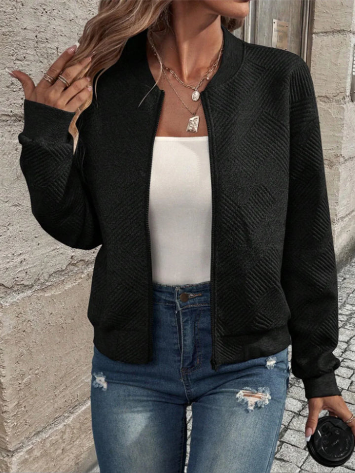 Woman wearing black textured bomber jacket, white tank top, distressed blue jeans, and layered gold necklaces