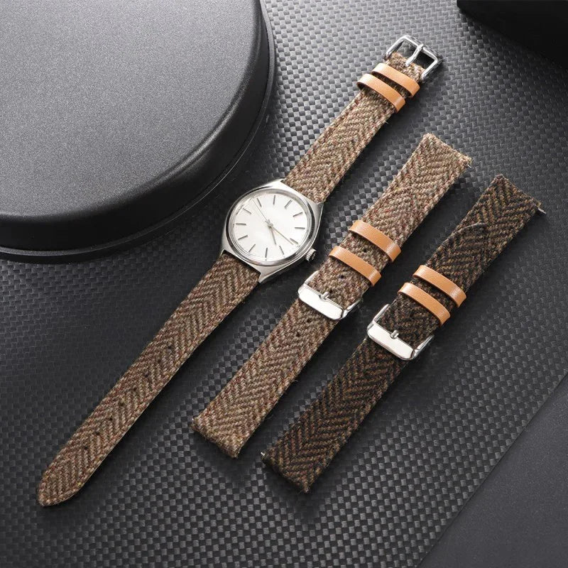 Silver watch with white dial and brown tweed fabric strap alongside two additional brown tweed straps on black textured surface