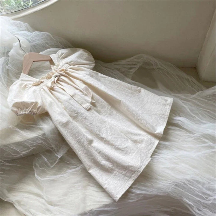 Cream-colored children's dress with puff sleeves and bow detail on wooden hanger on sheer fabric