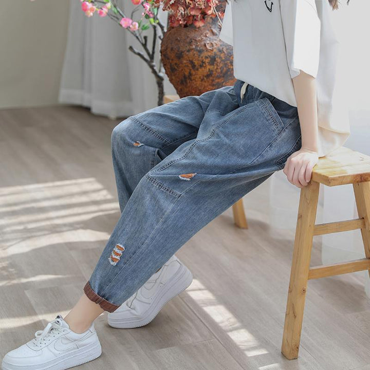 Casual denim jogger pants with distressed details worn with white sneakers, seated on wooden stool indoors