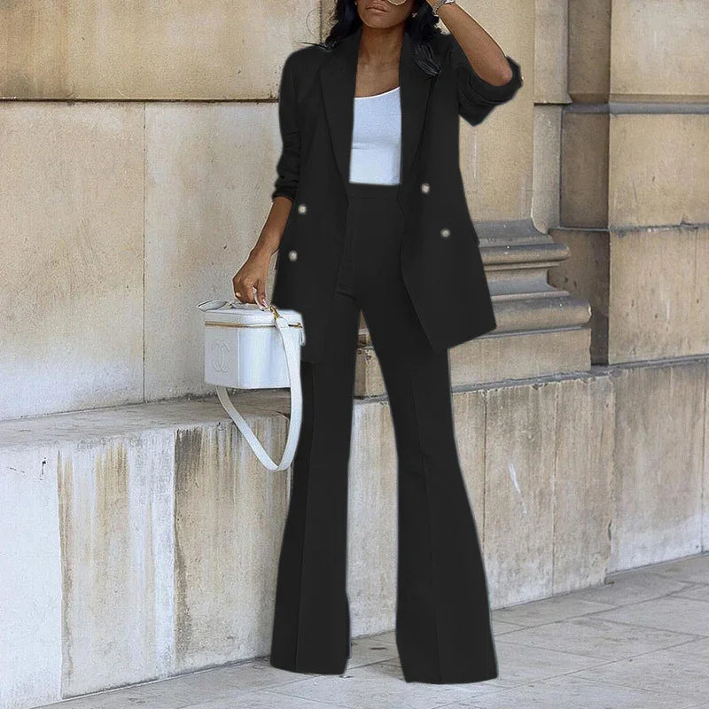 Woman wearing black blazer and flared pants with white top and handbag against stone wall