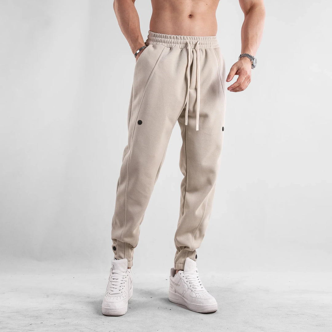 Man wearing beige jogger pants with black button details and white sneakers on grey background