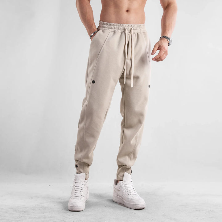 Man wearing beige jogger pants with black button details and white sneakers on grey background
