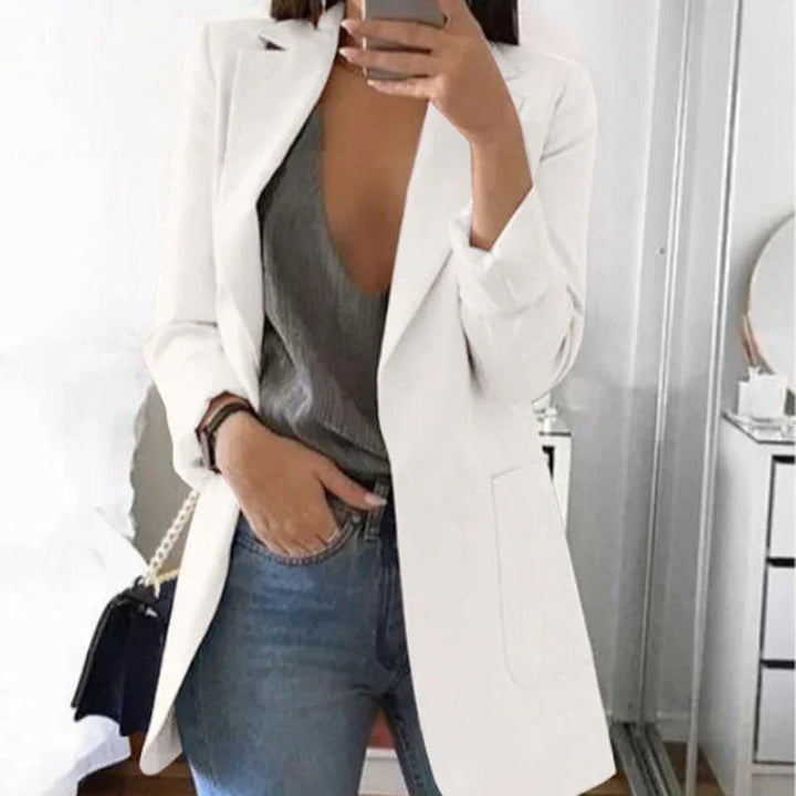 Woman wearing white blazer over gray top and blue jeans taking mirror selfie indoors