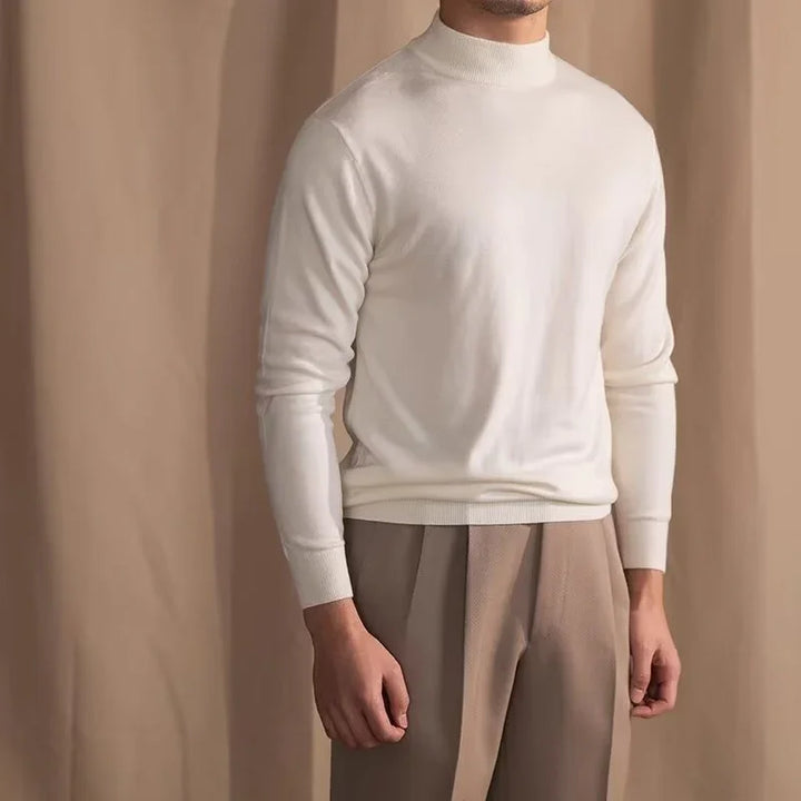 Man wearing white mock neck sweater and beige pleated trousers against neutral background