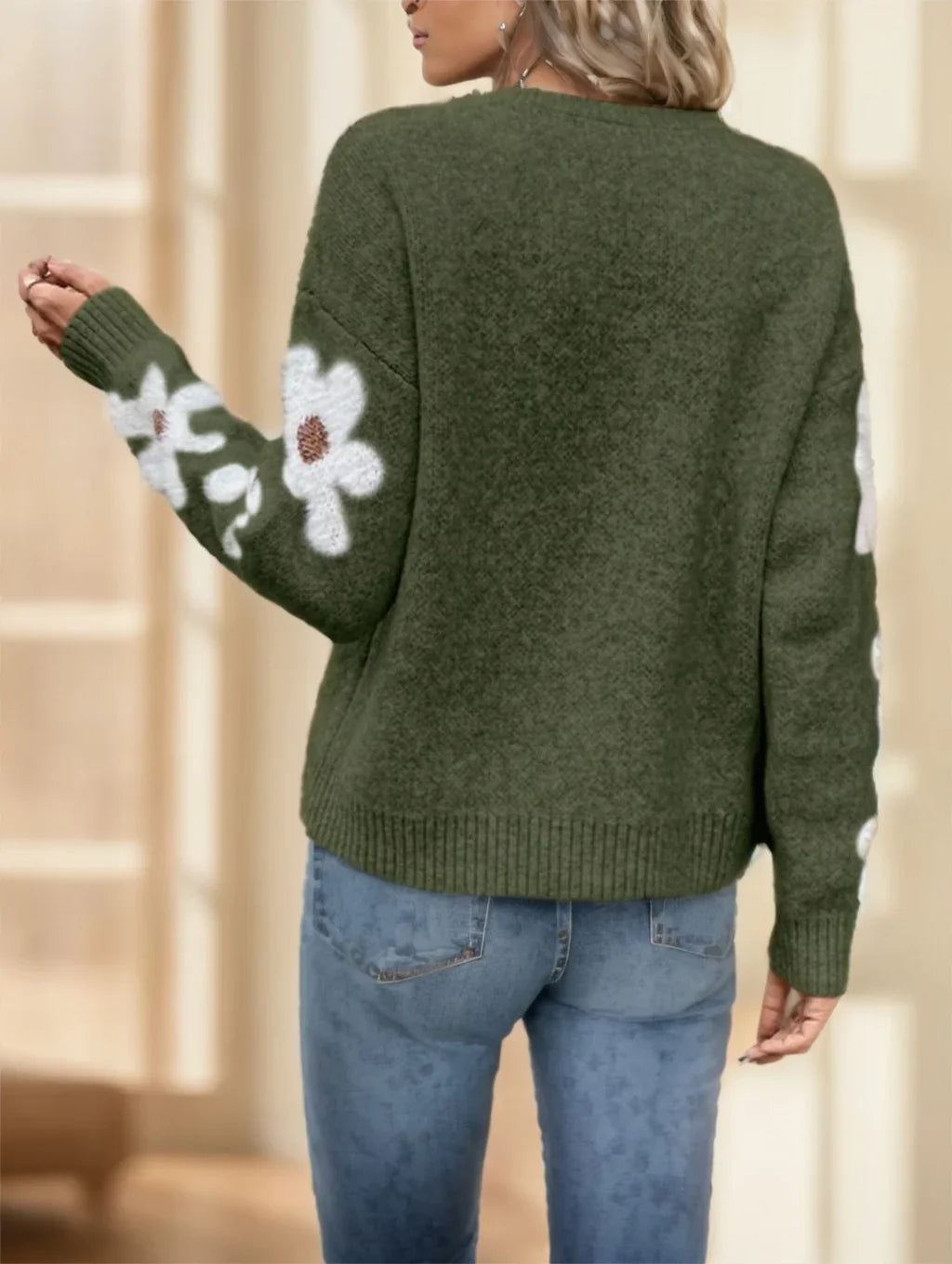 Woman wearing olive green sweater with white floral patterns on sleeves and blue jeans indoors