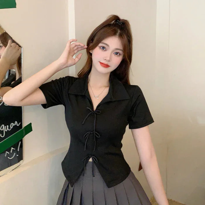 Young woman wearing black short-sleeve top with front bows and gray pleated skirt posing indoors