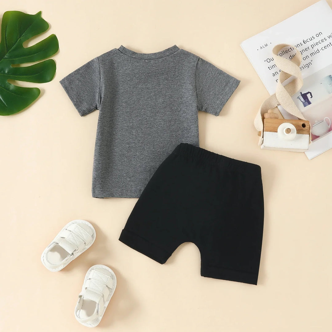 Baby outfit with grey short sleeve shirt, black shorts, white shoes, wooden toy camera on beige background