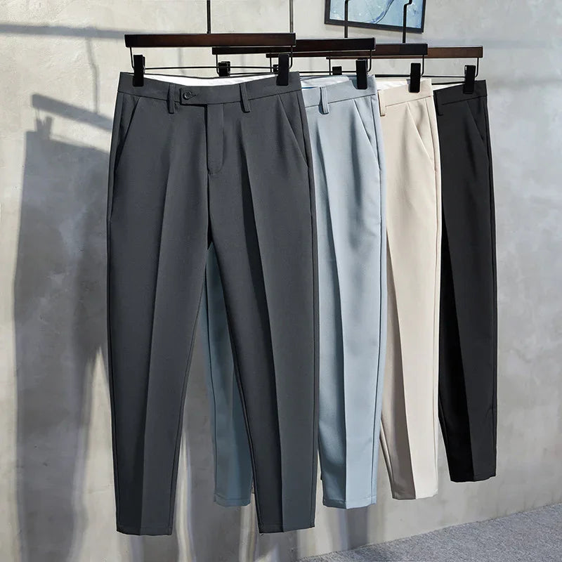 Four pairs of men's tailored trousers in grey, light blue, beige, and black hanging on wooden hangers in a minimalist setting