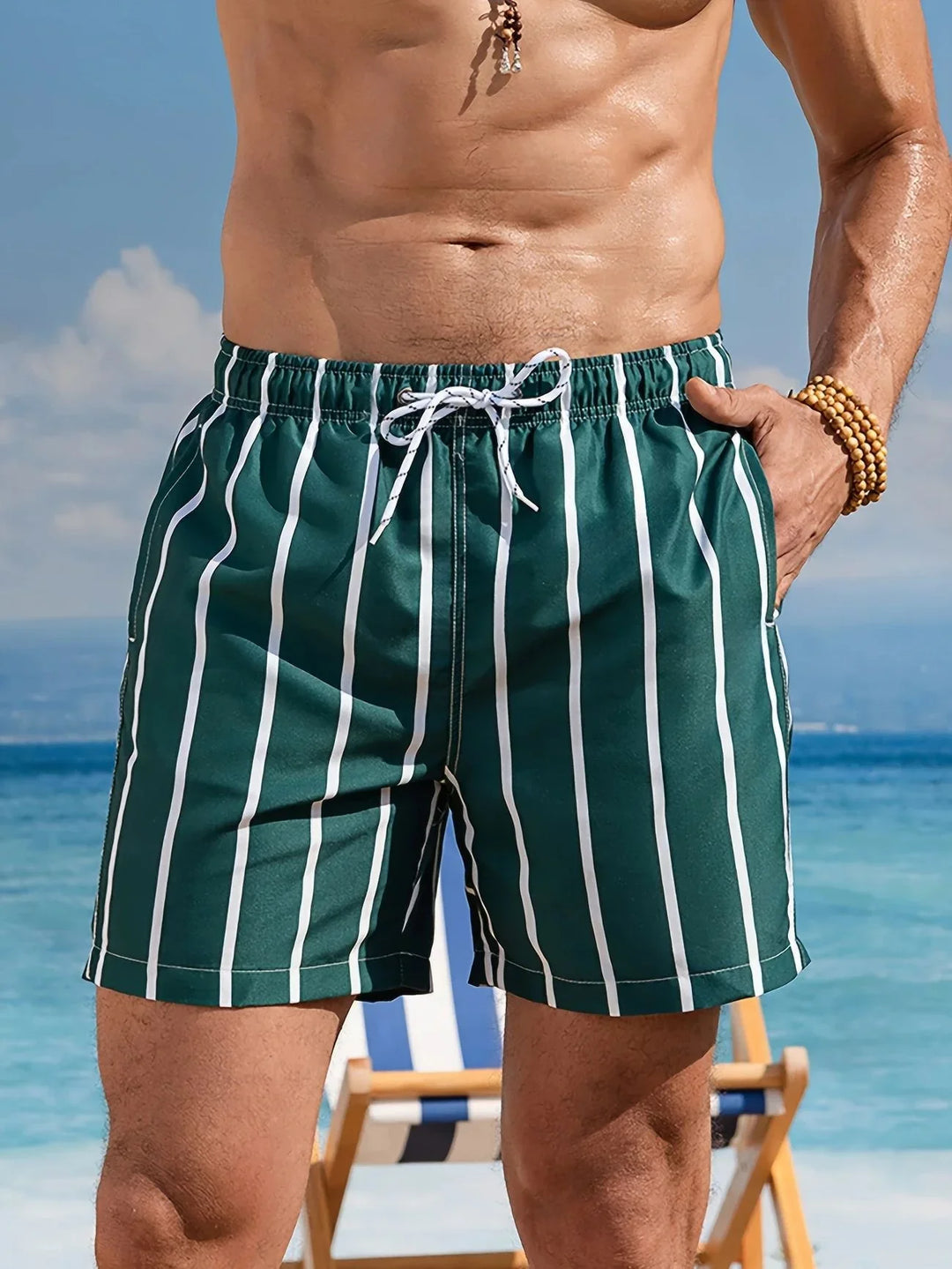 Man wearing green and white vertical striped swim trunks standing by the beach with a wooden chair in the background