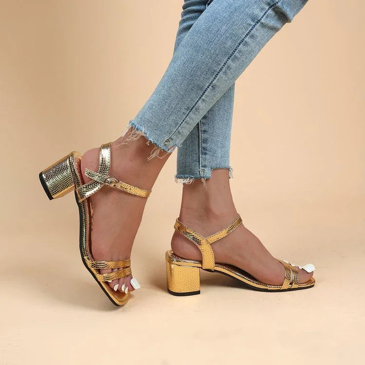 Women's feet wearing gold metallic textured block heel sandals with ankle straps and frayed hem blue jeans