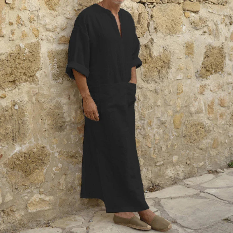 Man wearing a black traditional long robe and beige slip-on shoes leaning against a stone wall
