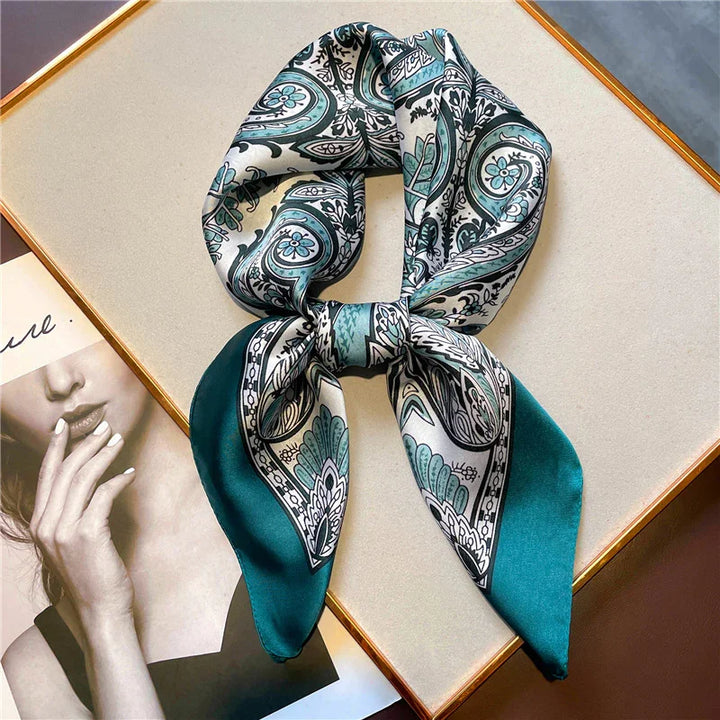 Silk scarf with teal, black, and white paisley pattern tied in a knot on beige surface next to fashion magazine