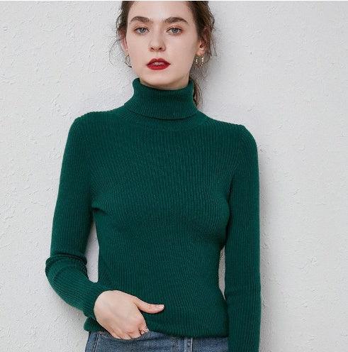 Young woman wearing a fitted dark green ribbed turtleneck sweater against a light textured wall