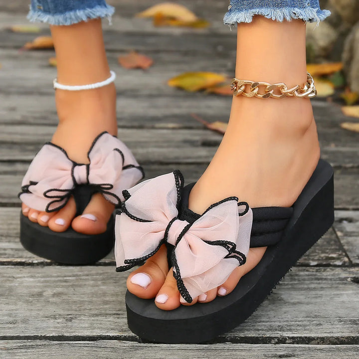 Wedge platform sandals with large pink bows and black trim on wooden deck, worn with frayed jeans and anklets
