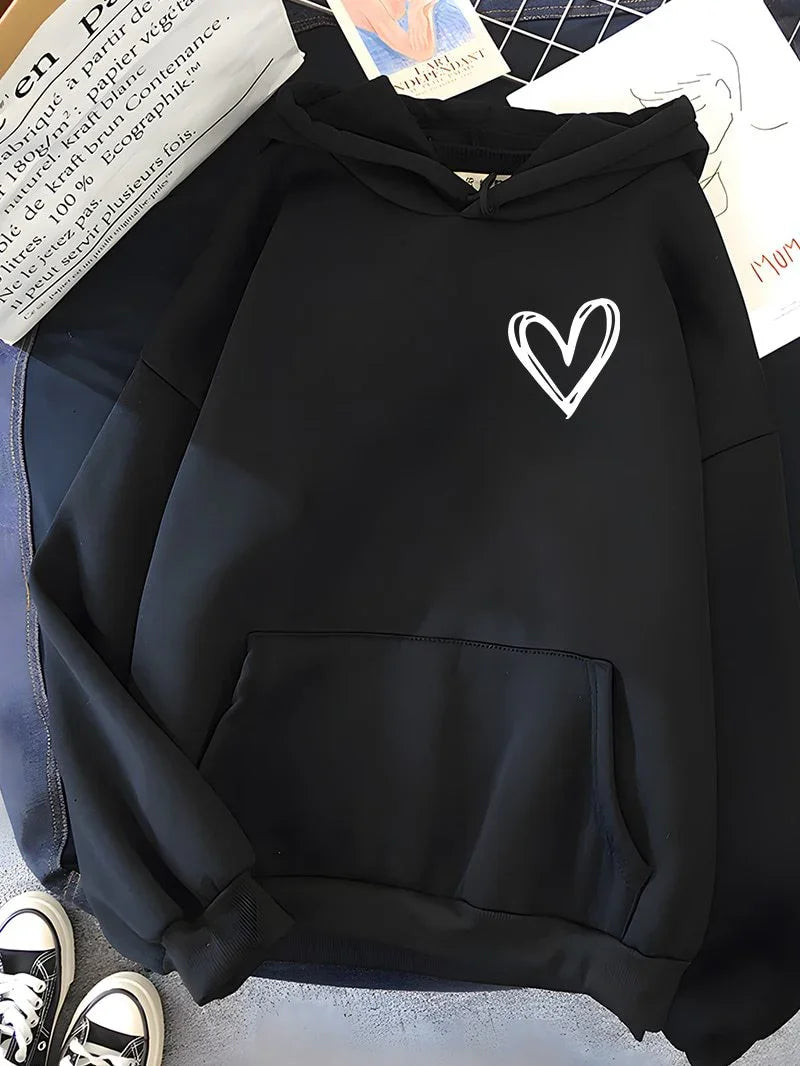 Black hoodie with a white hand-drawn heart on chest, laid flat on a blue surface with a pair of black and white sneakers nearby