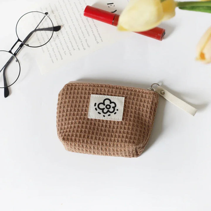 Brown textured fabric pouch with flower patch, black round glasses, red lipstick, and yellow tulips on white surface