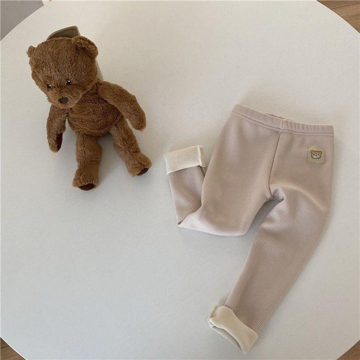 Brown teddy bear plush toy next to beige ribbed baby leggings with teddy patch on white round table