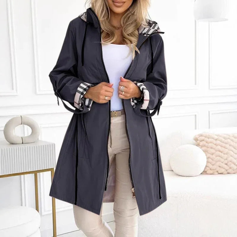 Woman wearing black hooded raincoat with plaid cuffs and beige pants in modern white living room