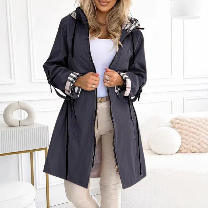 Woman wearing black hooded raincoat with plaid cuffs and beige pants in modern white living room