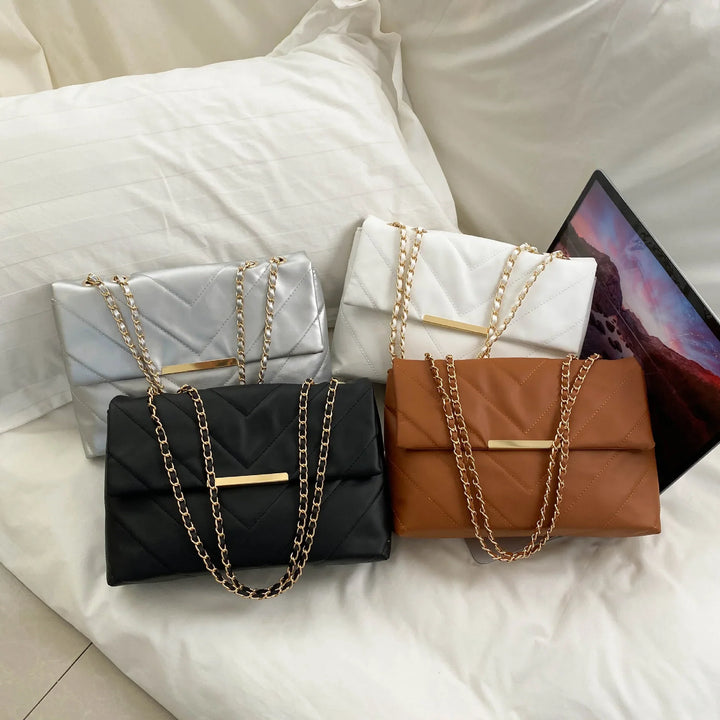 Four quilted leather chain strap handbags in silver, white, black, and brown on a white bed
