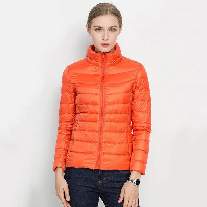 Woman wearing a bright orange quilted puffer jacket with high collar and dark jeans against white background