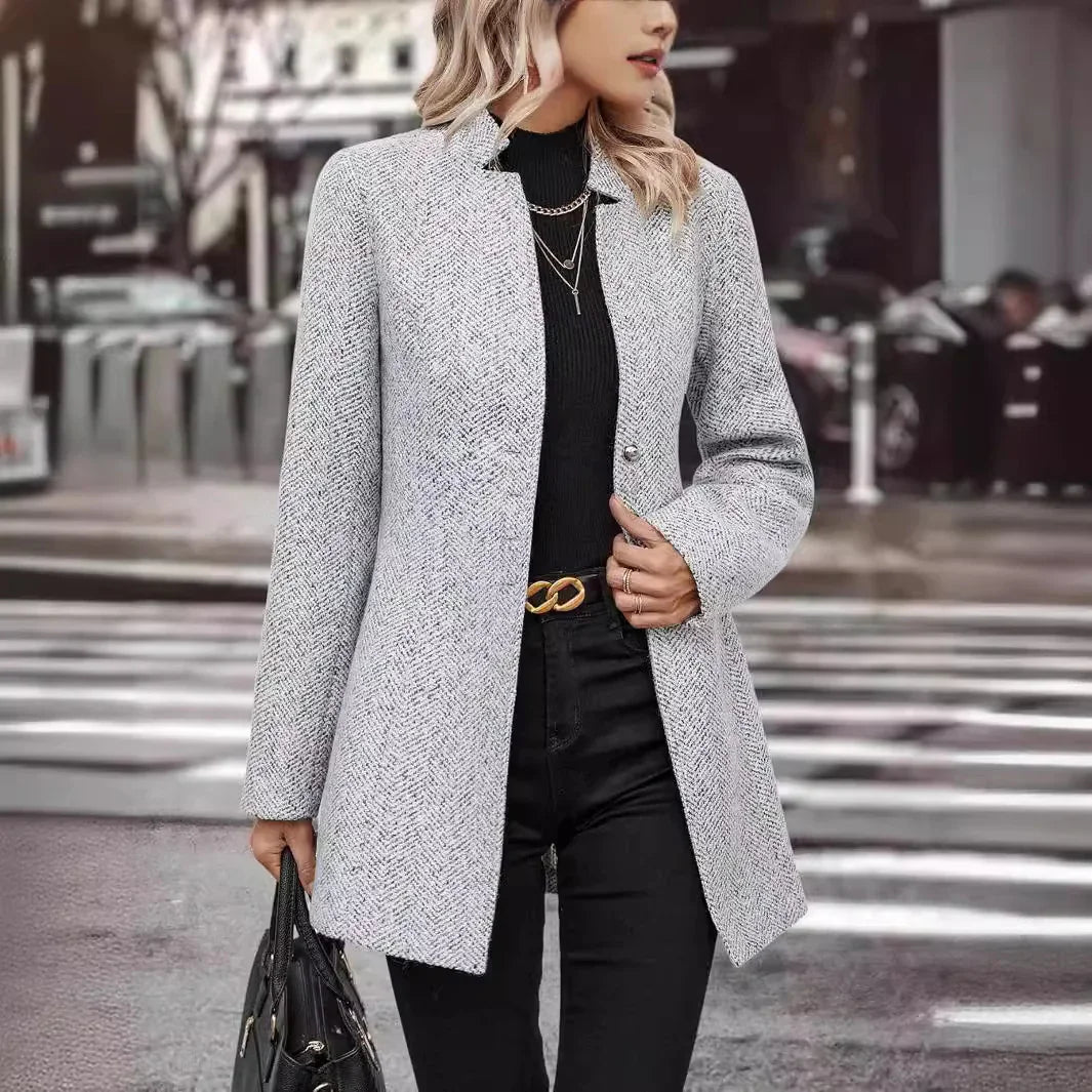 Woman wearing a stylish gray herringbone coat over a black turtleneck with layered necklaces in urban street setting
