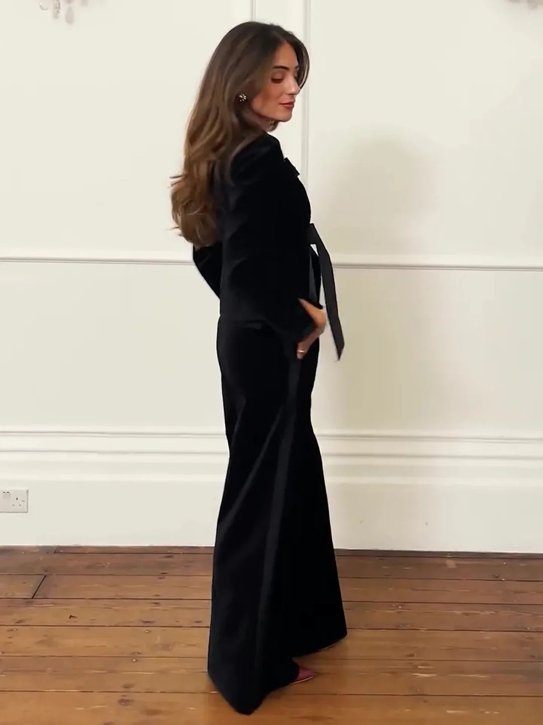 Woman in elegant black long dress with ribbon belt standing on wooden floor against white wall