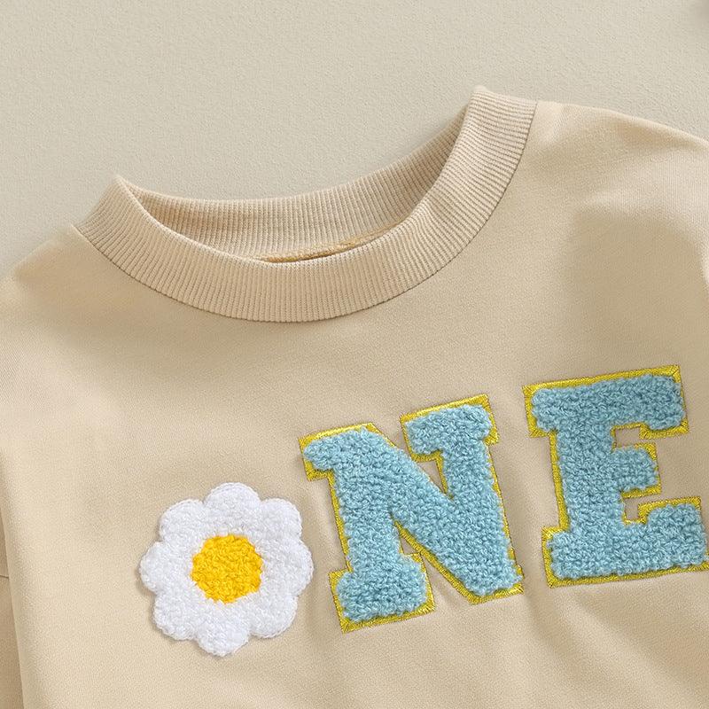 Beige sweatshirt with textured blue letters 'NE' and a white and yellow flower patch design