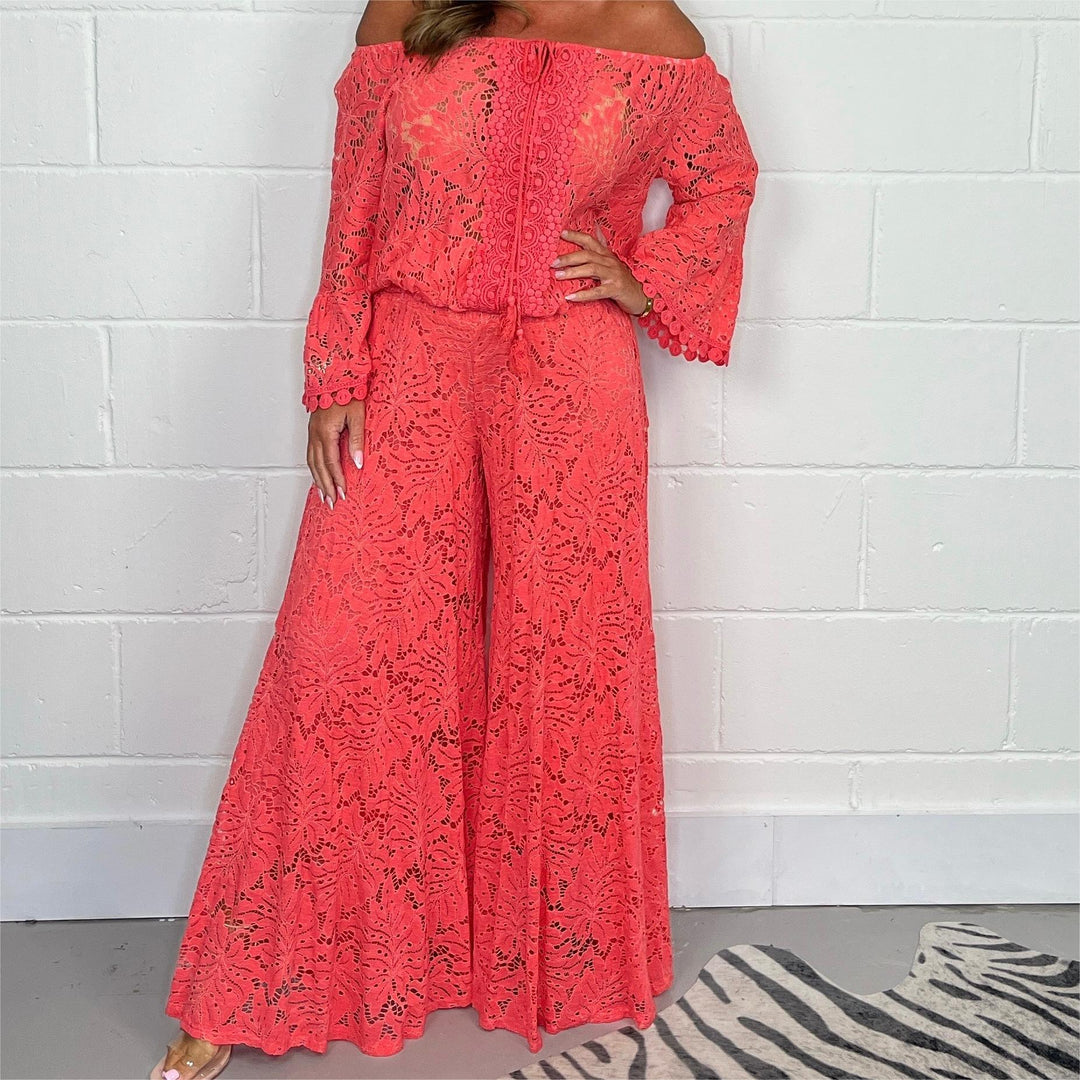 Woman wearing bright coral lace off-shoulder top and wide-leg pants against white brick wall