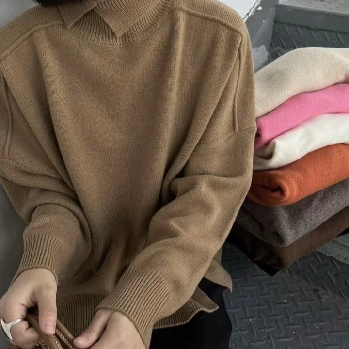 Person wearing brown turtleneck sweater with folded sweaters in pink, cream, orange, gray, and brown on side