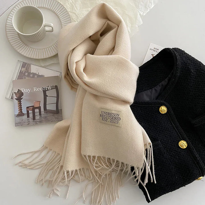Cream scarf with fringe and label 'UNDRESON REGISTERED EST. 2023' beside black textured jacket with gold buttons, white teacup and saucer, and CD case