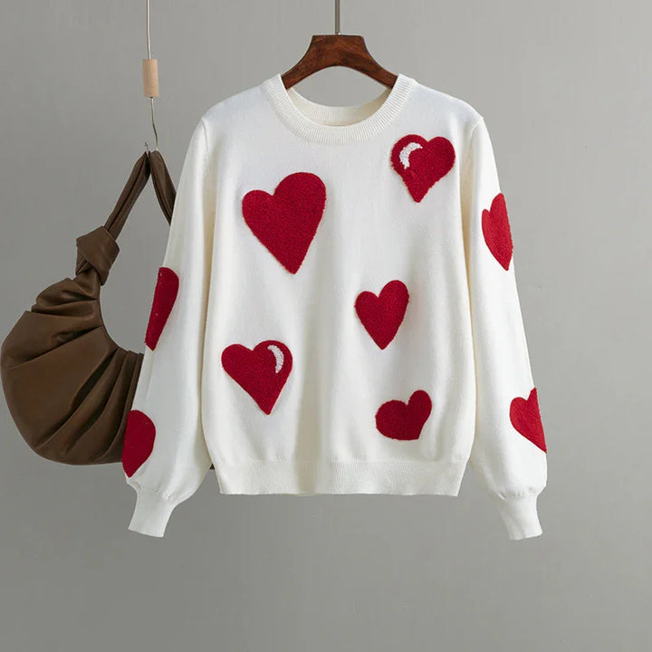 White sweater with red heart patterns on front and sleeves hanging next to brown handbag