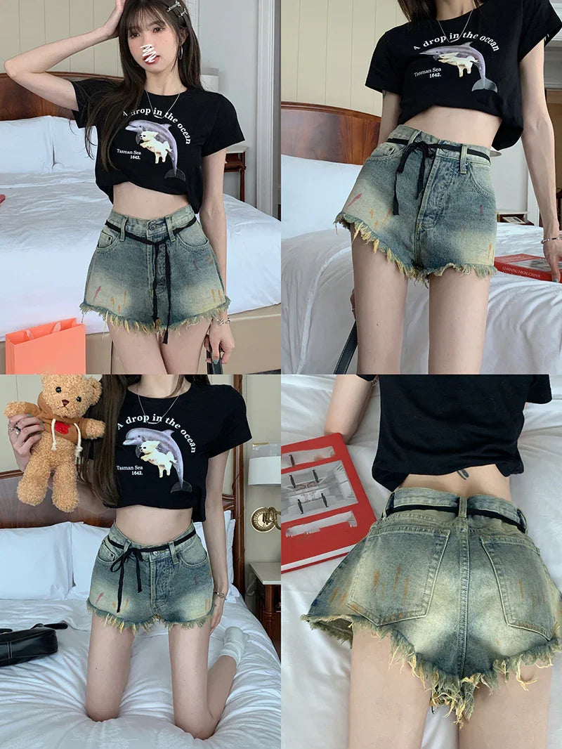 Young woman in black cropped dolphin graphic tee and distressed high-waist denim shorts with black belt in bedroom