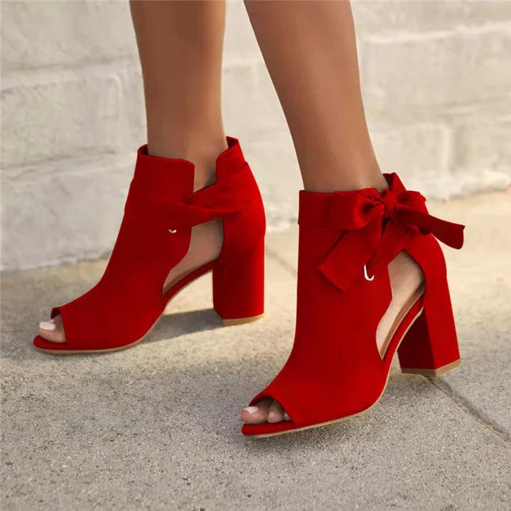 Red suede peep-toe block heel ankle boots with cutouts and decorative bow on sidewalk