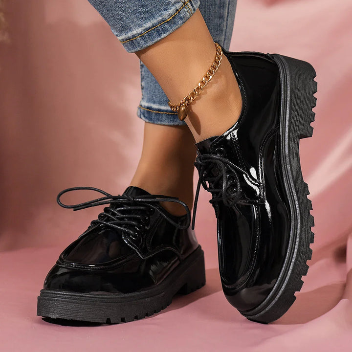 Close-up of stylish black patent leather platform shoes worn with rolled-up jeans and a gold anklet on a pink background