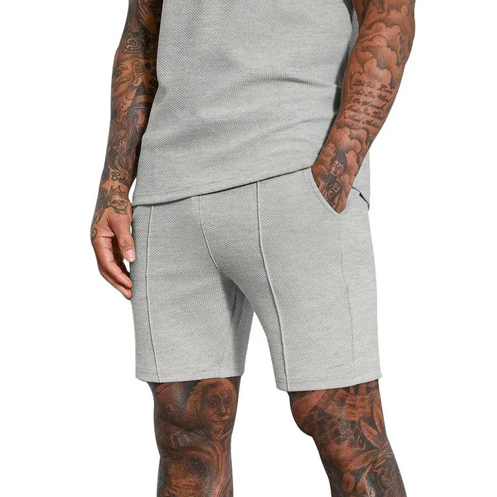 Close-up of man wearing light gray textured shorts and matching top with tattooed arms and legs